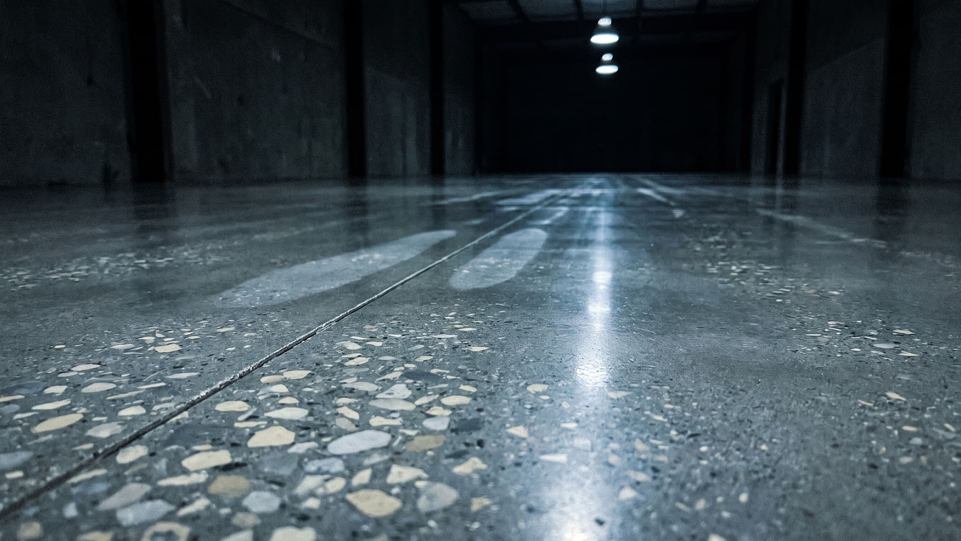 A mechanically polished concrete floor in a dark hall, pale exposed aggregate sharp in the foreground, a saw-cut control joint crossing the slab and pendant lights reflected down its length
